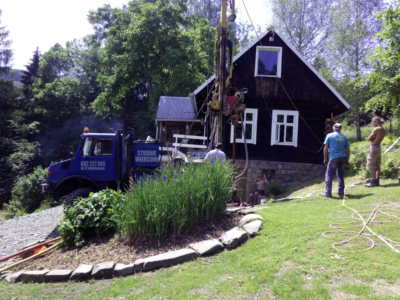 Wiercenie studni przy drewnianym domu na wzgórzu, widoczny specjalistyczny samochód z napisem 'Studnie Wiercone' i ekipa w trakcie pracy.