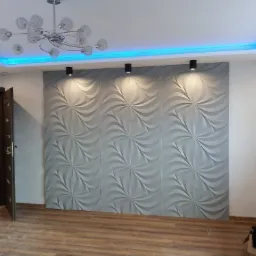 Ściana z paneli 3D w kolorze szarym, oświetlona trzema punktowymi lampami sufitowymi i podświetleniem LED w kolorze niebieskim wokół sufitu podwieszanego, widoczna z parkietem na podłodze...