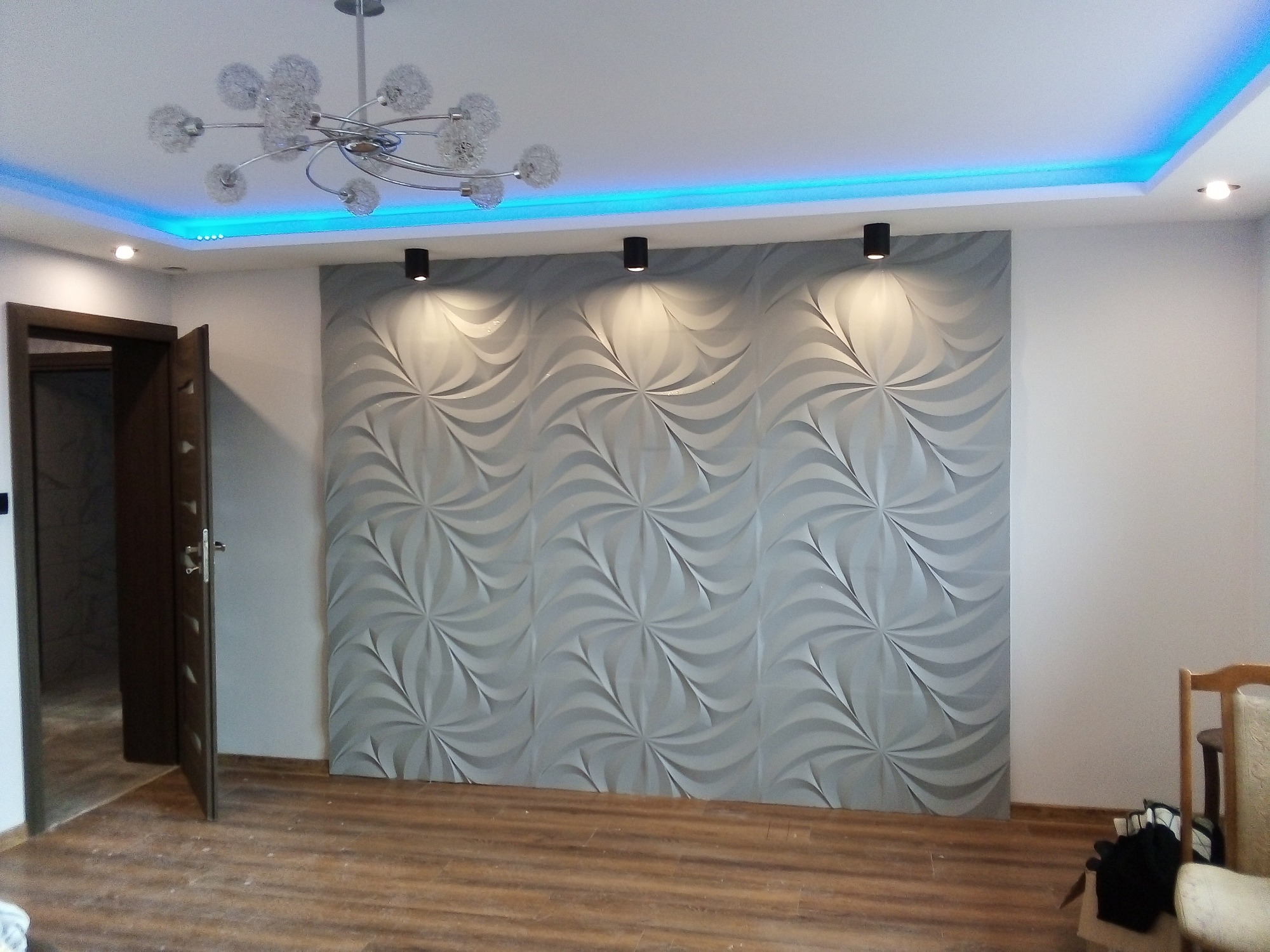 Ściana z paneli 3D w kolorze szarym, oświetlona trzema punktowymi lampami sufitowymi i podświetleniem LED w kolorze niebieskim wokół sufitu podwieszanego, widoczna z parkietem na podłodze...