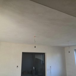 SF-Wykończenia Wnętrz (Quality) - Wnętrze pokoju w trakcie prac wykończeniowych, ściany pokryte gładzią, widoczne okno i drzwi balkonowe, sufit przygotowany do malowania, wiszące przewody elektryczne z żarówkami.