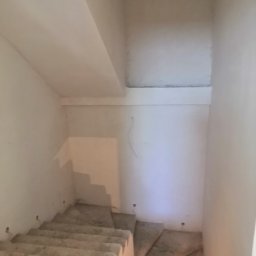 SF-Wykończenia Wnętrz (Quality) - Klatka schodowa w trakcie wykańczania: surowe, betonowe schody prowadzą w dół, ściany i sufit pokryte świeżym tynkiem w odcieniach bieli.