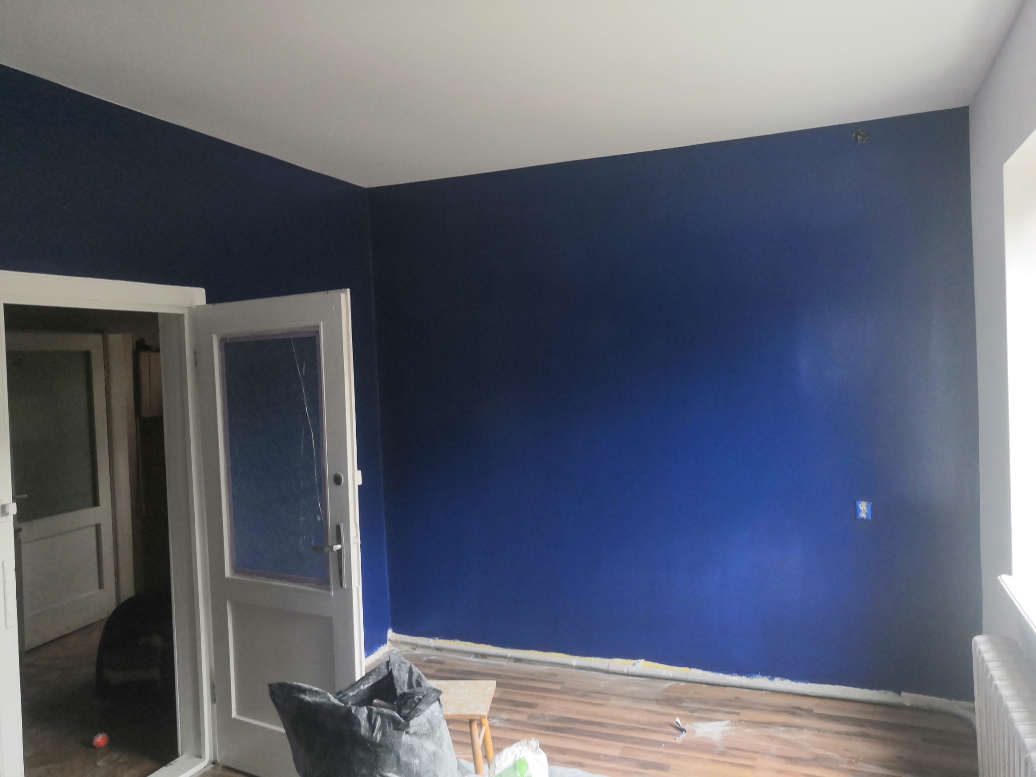 Pokój po malowaniu na ciemny odcień niebieskiego, widoczne ślady po taśmie malarskiej przy podłodze, otwarte białe drzwi i zabezpieczone gniazdko elektryczne.