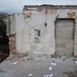 Częściowo rozebrana ściana budynku z odsłoniętą cegłą, resztkami tynku i widocznymi przewodami. Na podłodze gruz i fragmenty izolacji. W ścianie otwór zamurowany bloczkami.