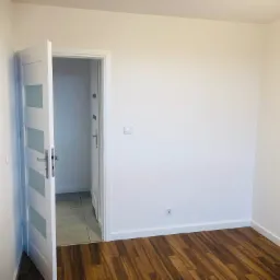 Pusty pokój po odświeżeniu: białe ściany, brązowe panele podłogowe, otwarte białe drzwi z czterema mlecznymi szybami, widoczny fragment podłogi w przyległym pomieszczeniu.