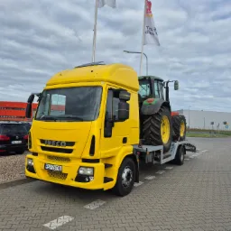 Żółta laweta Iveco z zielonym traktorem na platformie, zaparkowana na parkingu z kostki. W tle widoczne budynki i zaparkowany czarny van.