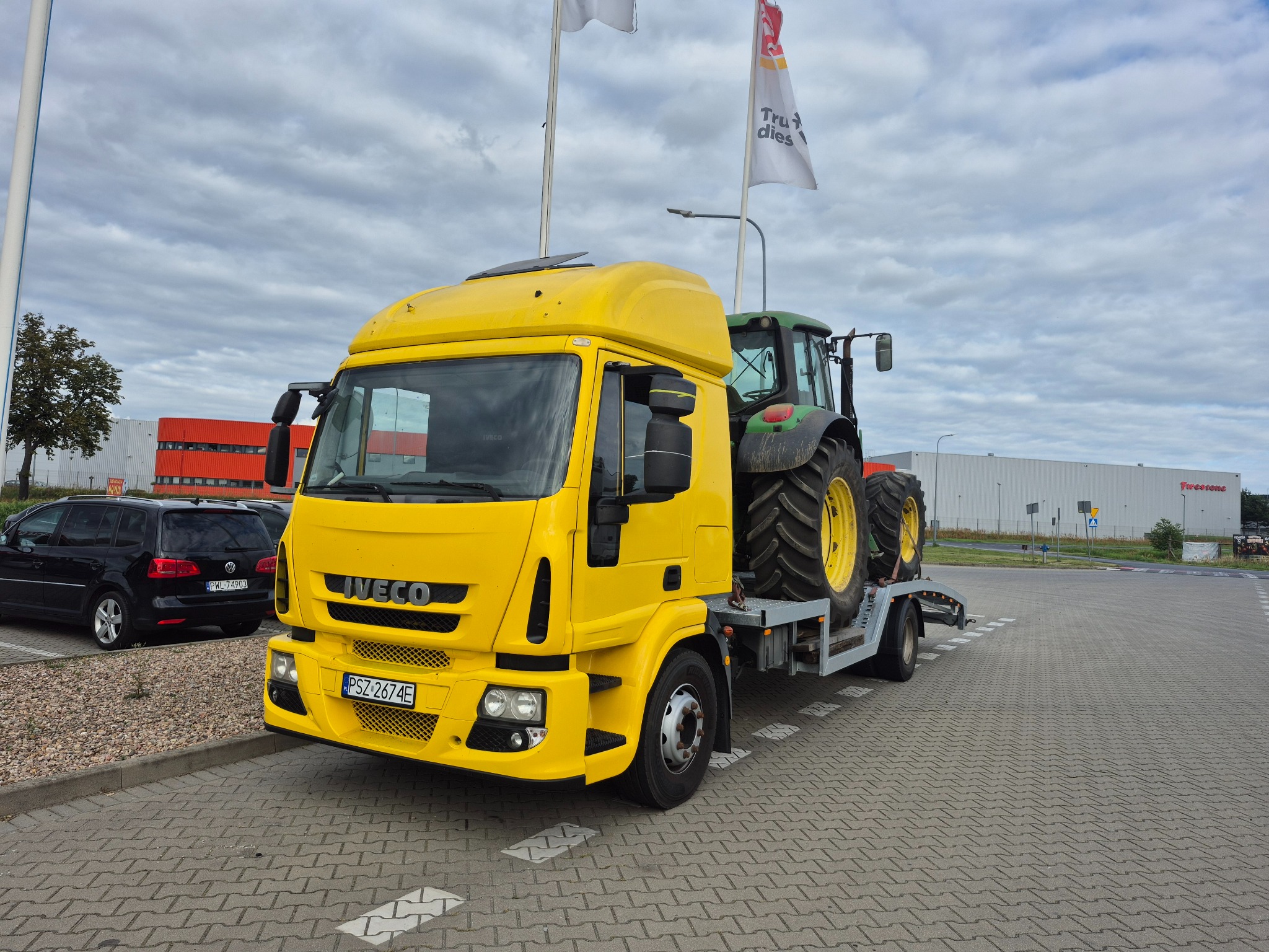 Żółta laweta Iveco z zielonym traktorem na platformie, zaparkowana na parkingu z kostki. W tle widoczne budynki i zaparkowany czarny van.