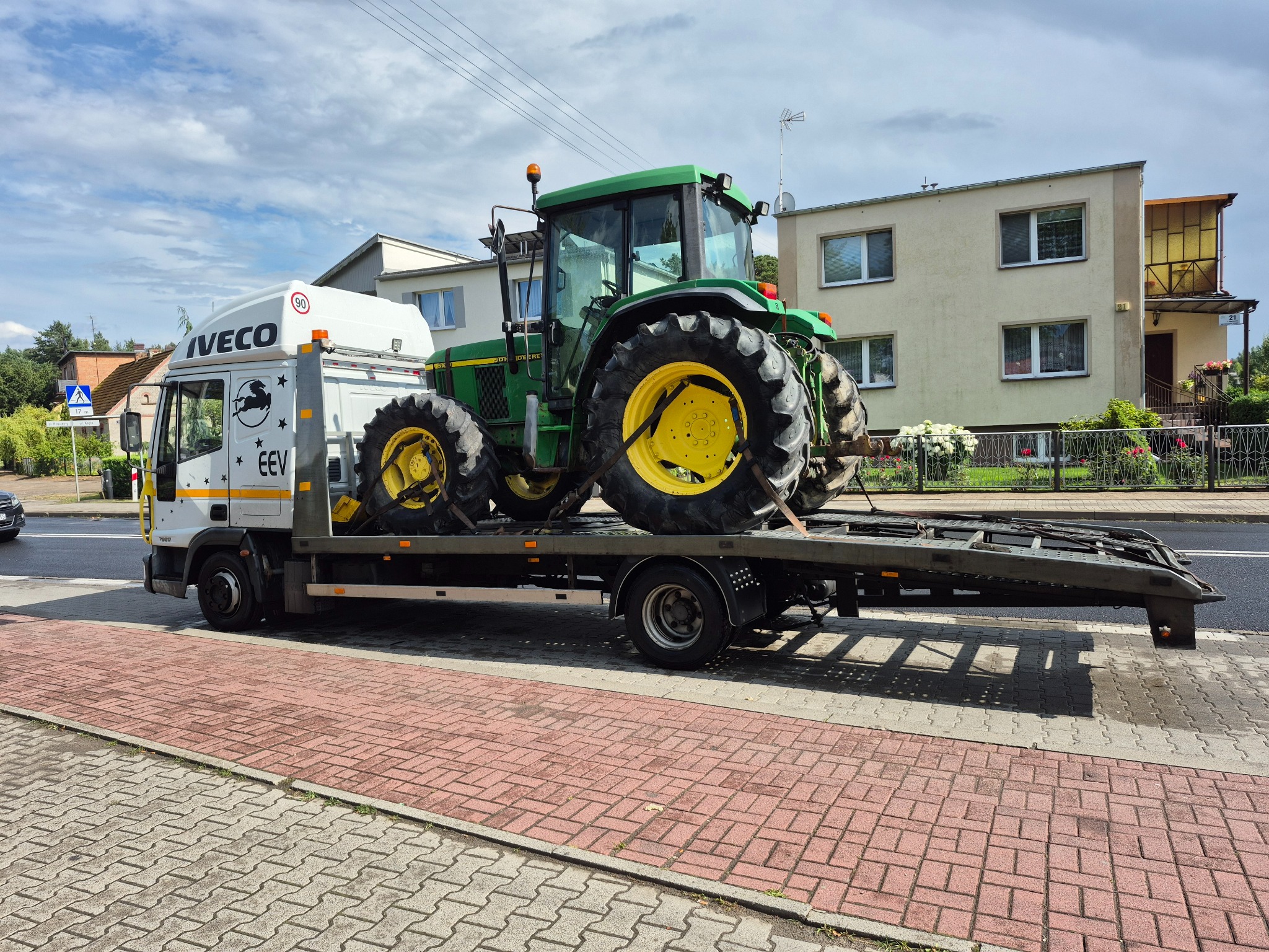 Laweta Iveco przewozi zielony traktor John Deere na tle budynków. Widoczne detale: tablica rejestracyjna EEV, znak ograniczenia prędkości 90 na kabinie.