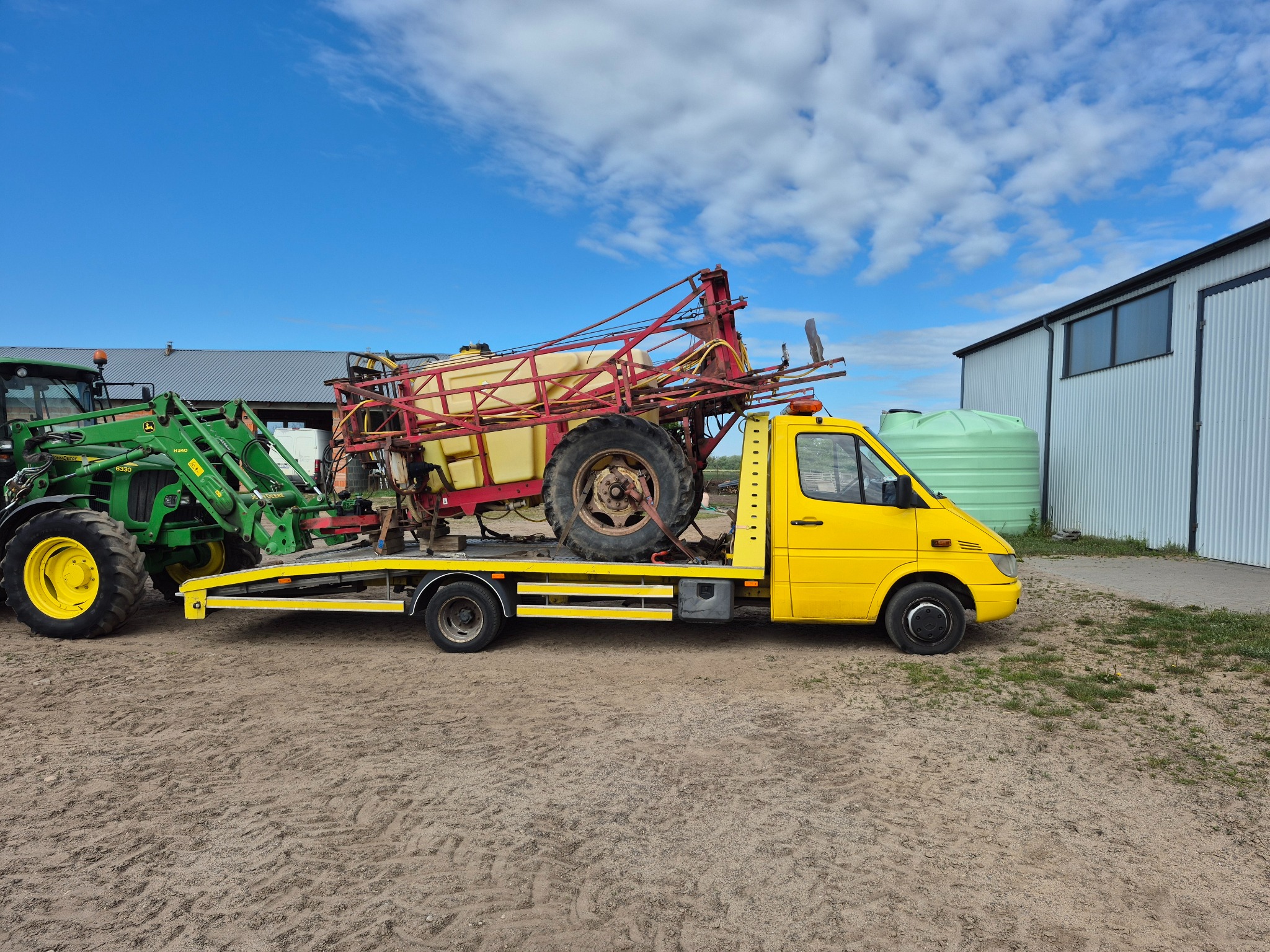 Żółta laweta z zielonym traktorem i opryskiwaczem na platformie, zaparkowana na piaszczystym terenie pod błękitnym niebem z chmurami.