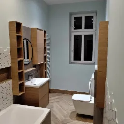 Jasna łazienka po remoncie z heksagonalnymi płytkami, drewnianymi szafkami, umywalką nablatową i podłogą ułożoną w jodełkę; widoczna pralka.