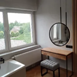 Jasna łazienka z drewnianymi akcentami: okrągłe lustro wiszące nad toaletką z jasnego drewna, szara pufa, biała wanna pod oknem z widokiem na zieleń, dekoracyjny panel ścienny z drewnianych listew.