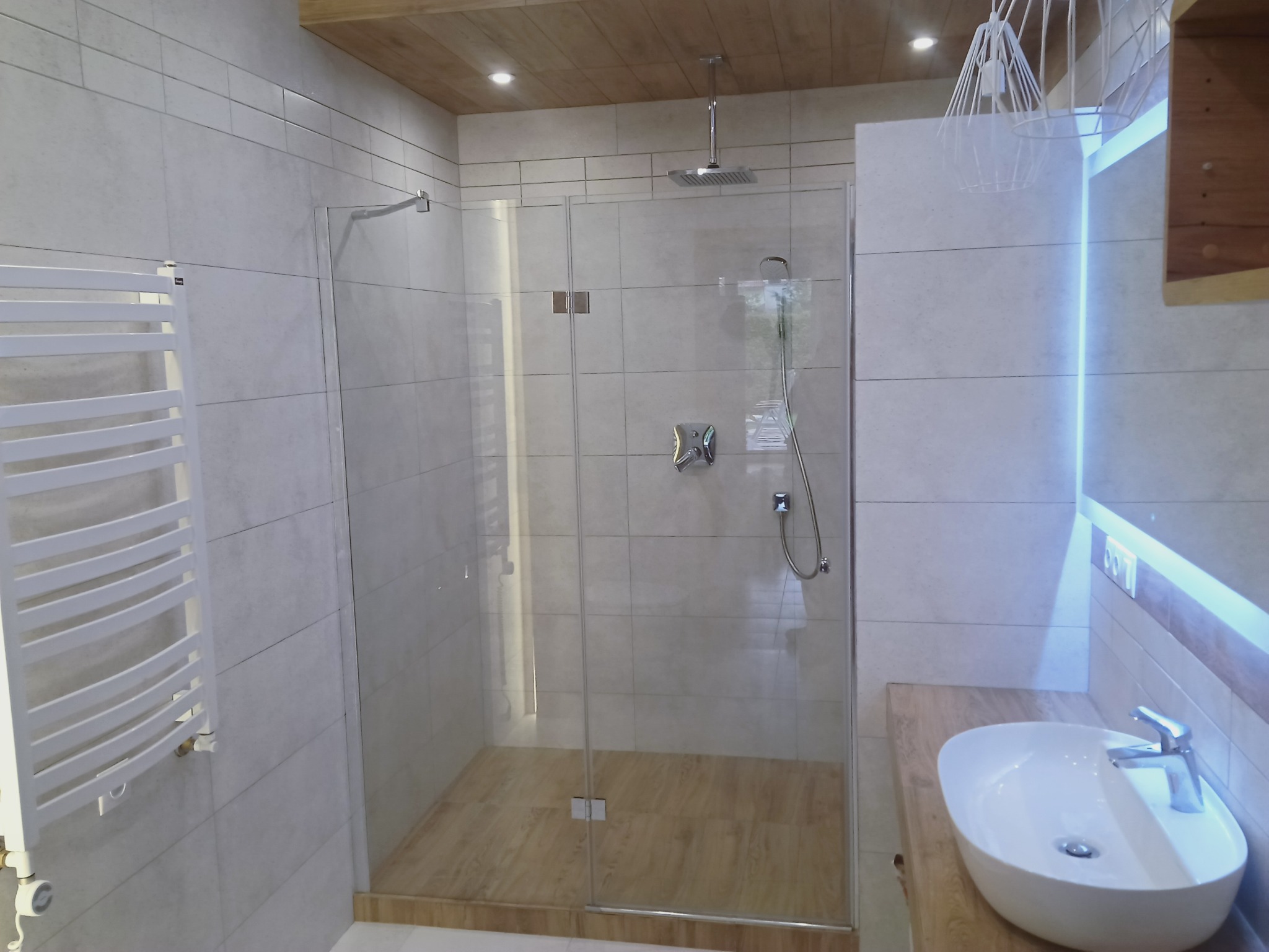 Nowoczesna łazienka z kabiną prysznicową typu walk-in, drewnianym sufitem i grzejnikiem drabinkowym. Umywalka nablatowa z podświetleniem LED.