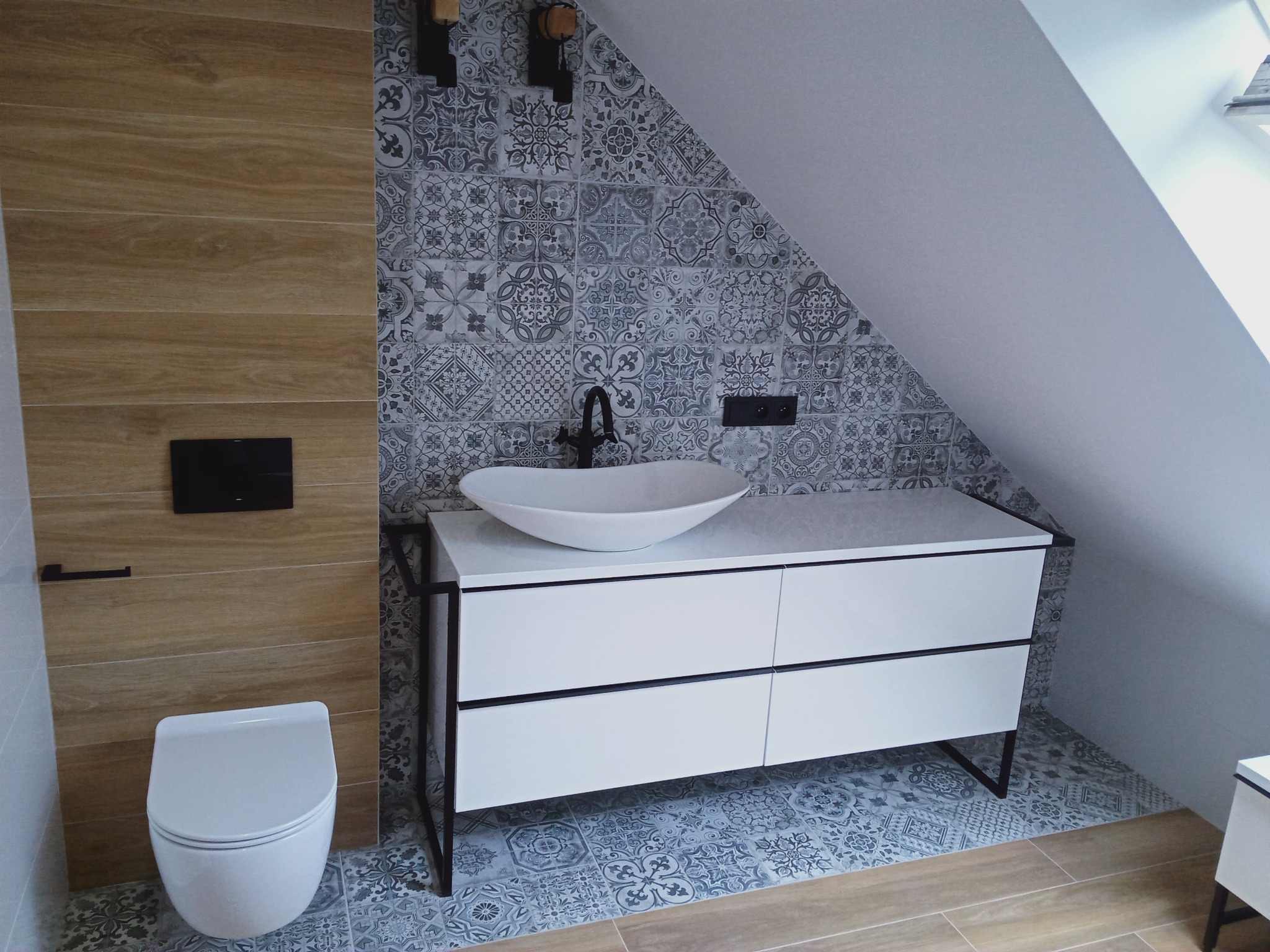 Nowoczesna łazienka na poddaszu z drewnopodobnymi i wzorzystymi płytkami, białą umywalką nablatową na szafce z czarnymi elementami oraz podwieszaną toaletą.