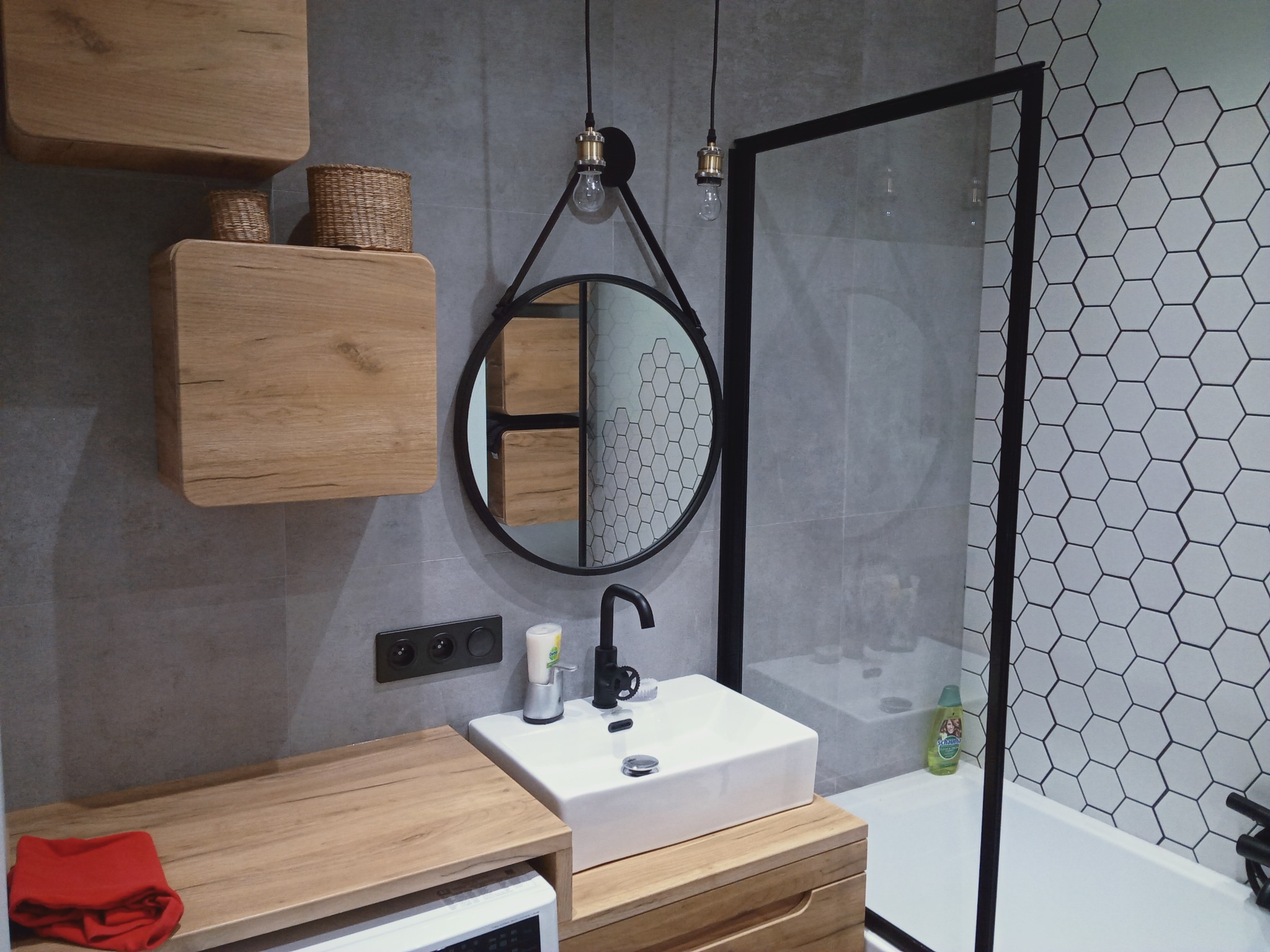 Nowoczesna łazienka z drewnianymi szafkami, okrągłym lustrem na pasku, czarną armaturą i heksagonalnymi płytkami za szklaną kabiną prysznicową.