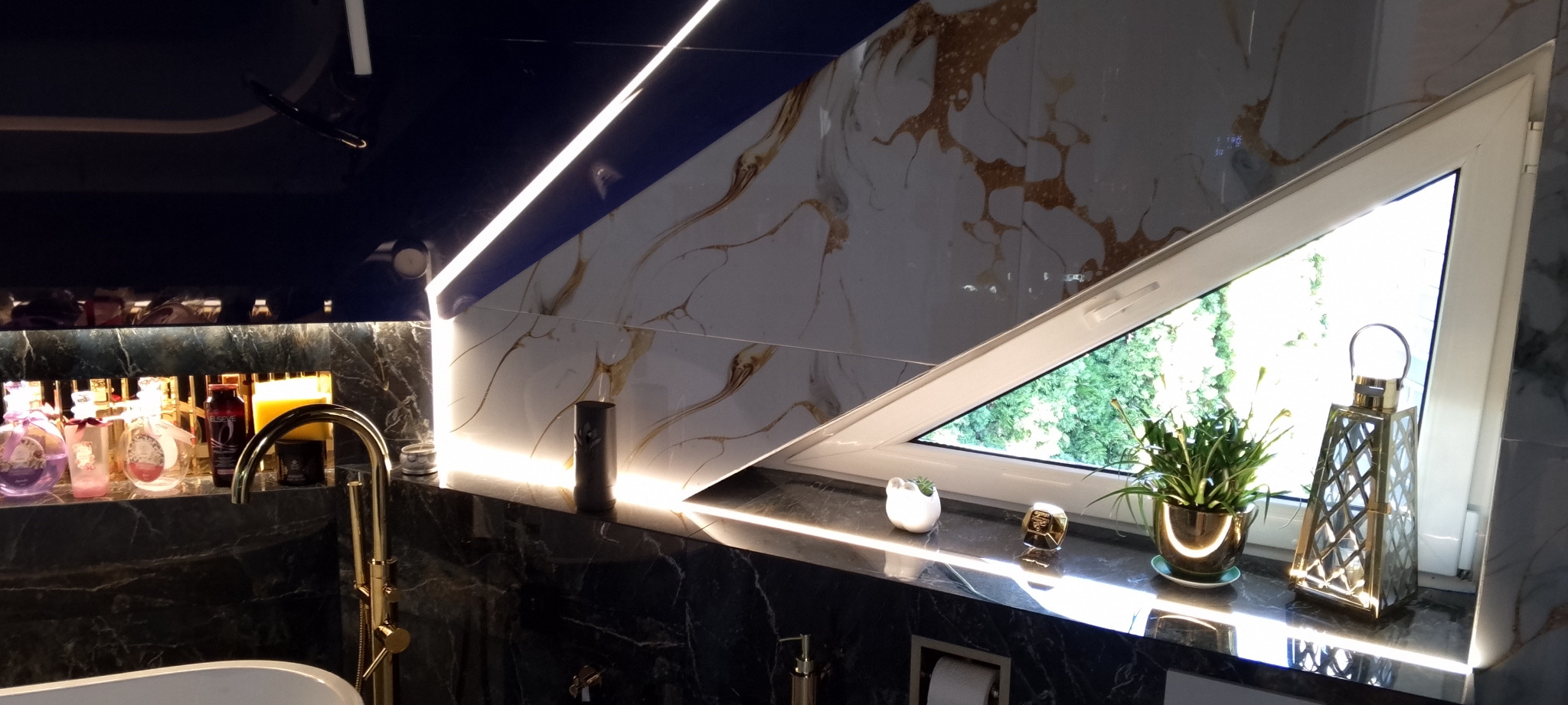 Luksusowa łazienka z marmurowymi płytkami, złotą armaturą i oknem w kształcie trapezu, oświetlona listwami LED. Widoczne kosmetyki i dekoracje.