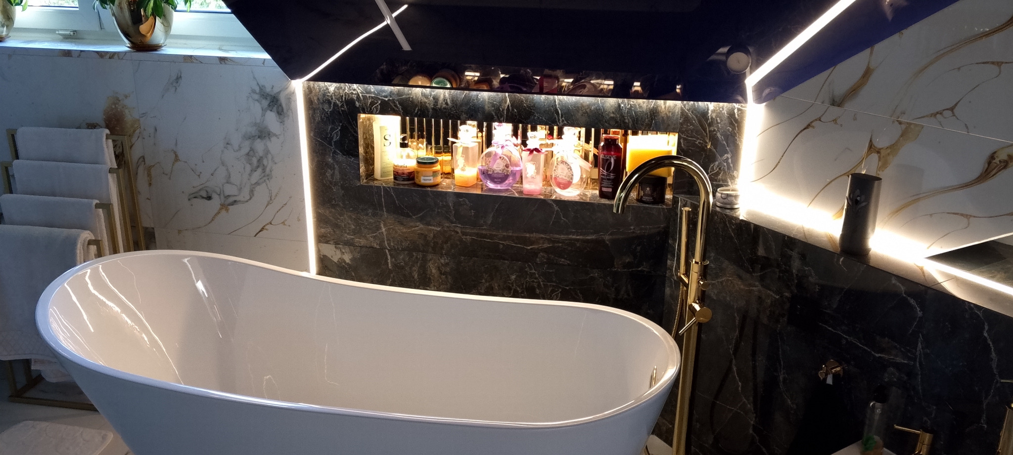 Elegancka łazienka z wolnostojącą wanną, marmurowymi płytkami i złotą armaturą. Wnęka z oświetleniem LED eksponuje kosmetyki. Nowoczesny design.