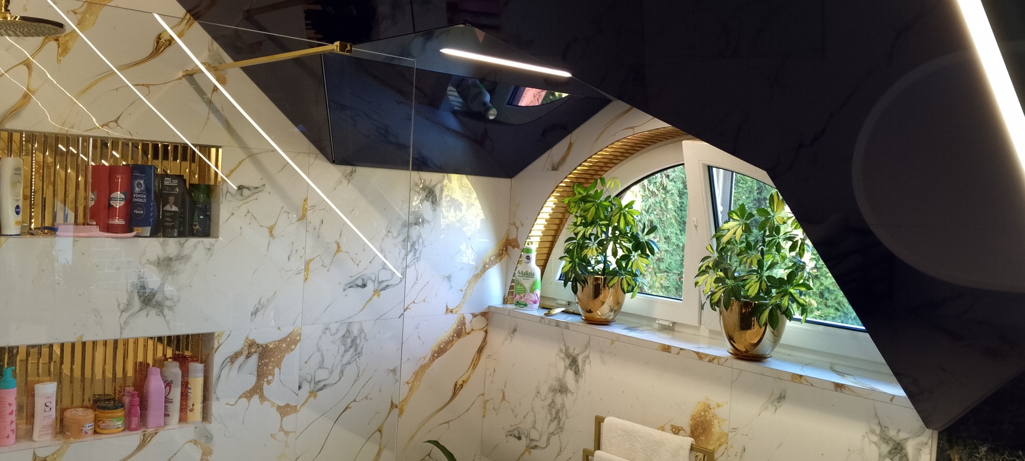 Elegancka łazienka z marmurowymi płytkami, złotymi akcentami i roślinami w doniczkach przy oknie. Widoczne półki z kosmetykami i kabina prysznicowa.