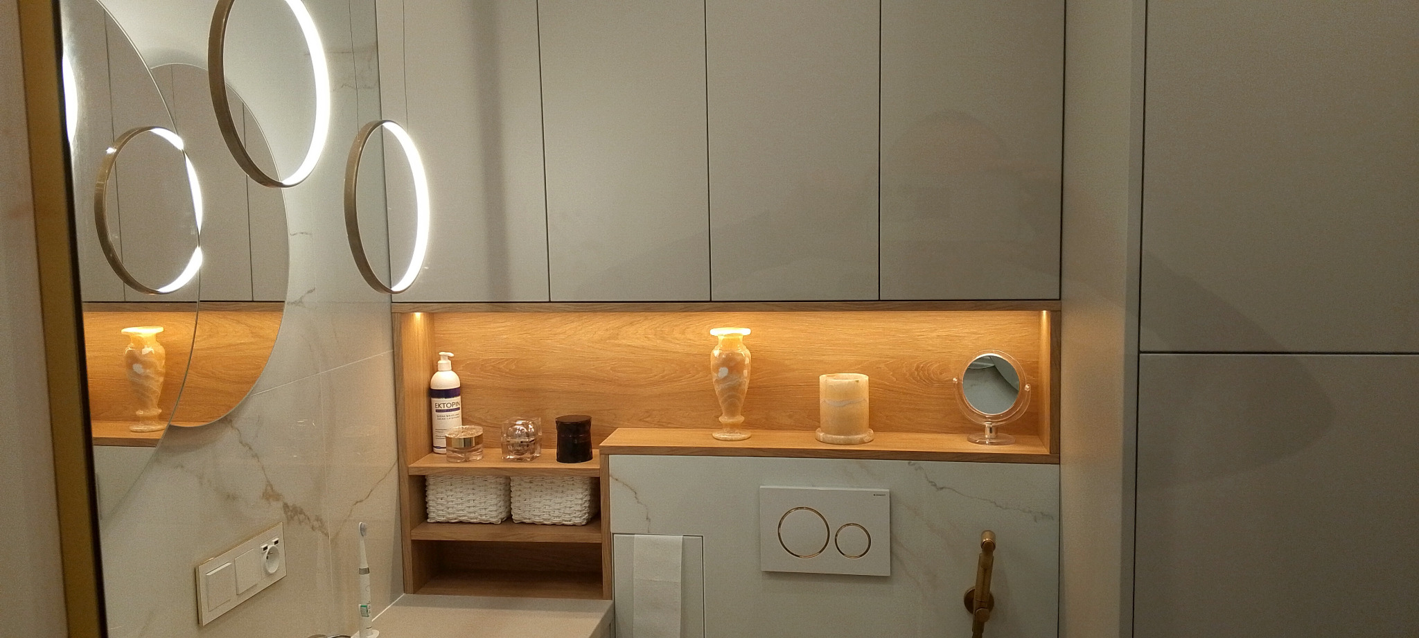 Nowoczesna łazienka z podświetlanymi okrągłymi lustrami, drewnianą wnęką z dekoracjami i szarą zabudową meblową. Marmurowe płytki na ścianach.