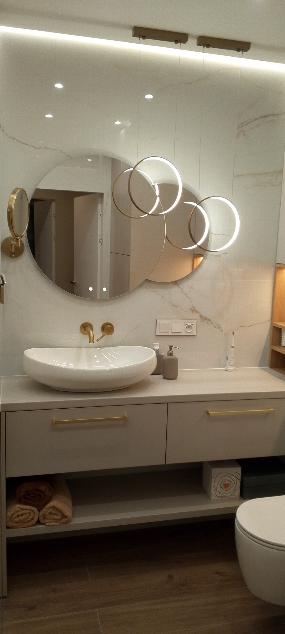 Nowoczesna łazienka z okrągłym lustrem z podświetleniem LED, białą umywalką nablatową i złotą armaturą. Marmurowe płytki na ścianach i drewnopodobna podłoga.