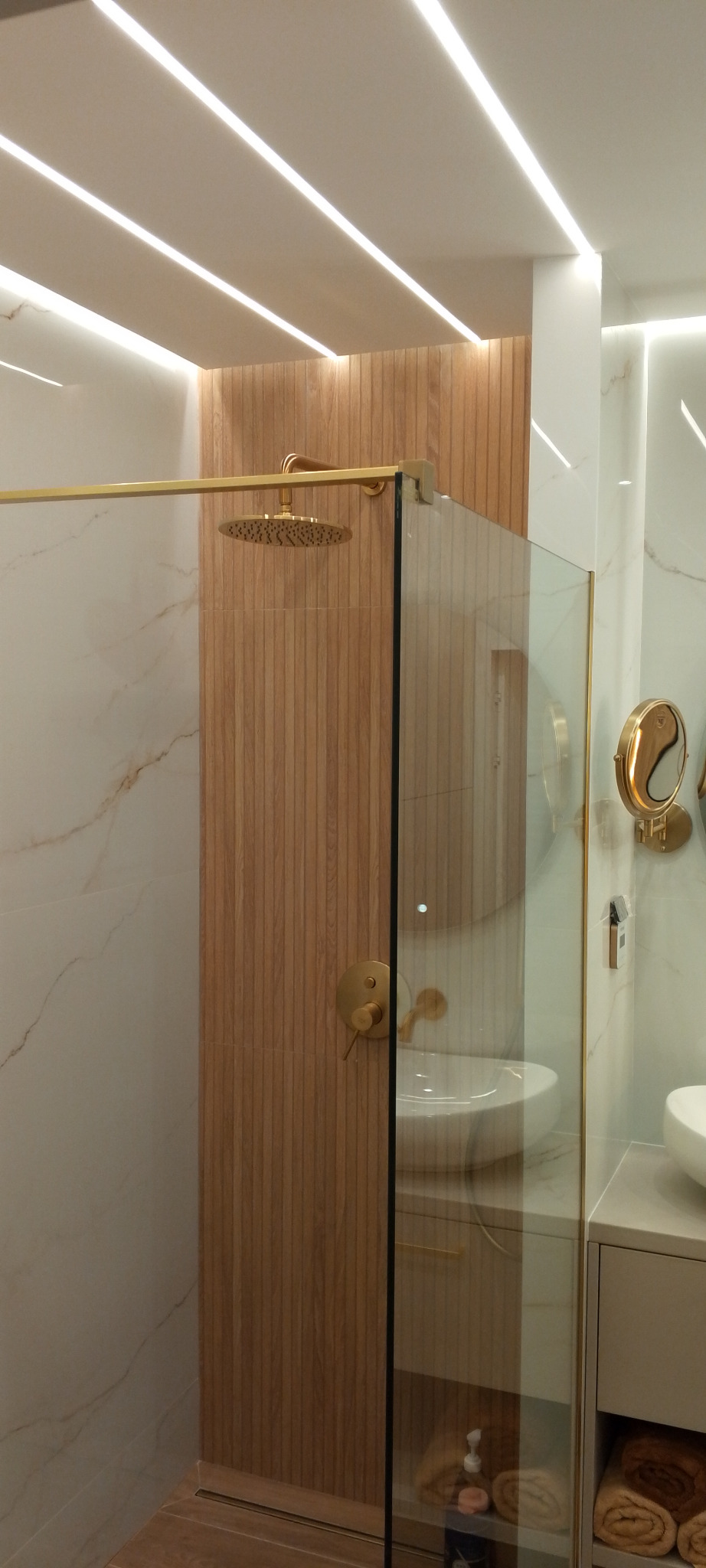 Nowoczesna kabina prysznicowa z deszczownicą w odcieniach bieli, beżu i złota. Drewnopodobne panele ścienne i marmurowe płytki. Oświetlenie LED podkreśla minimalistyczny design.