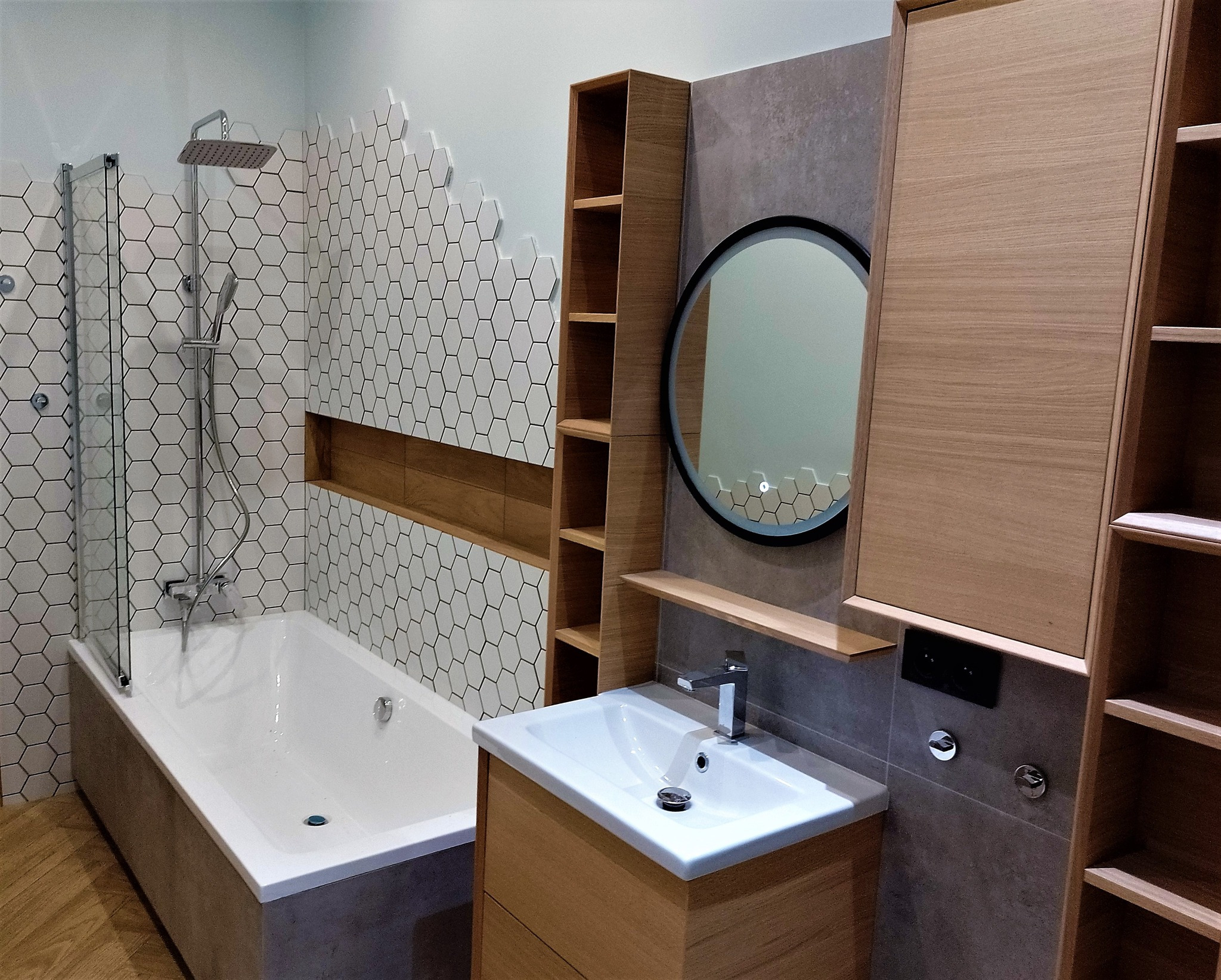 Nowoczesna łazienka z wanną i kabiną prysznicową za szklaną ścianką, umywalką w drewnianej szafce, okrągłym lustrem z podświetleniem LED i heksagonalnymi płytkami na ścianie.