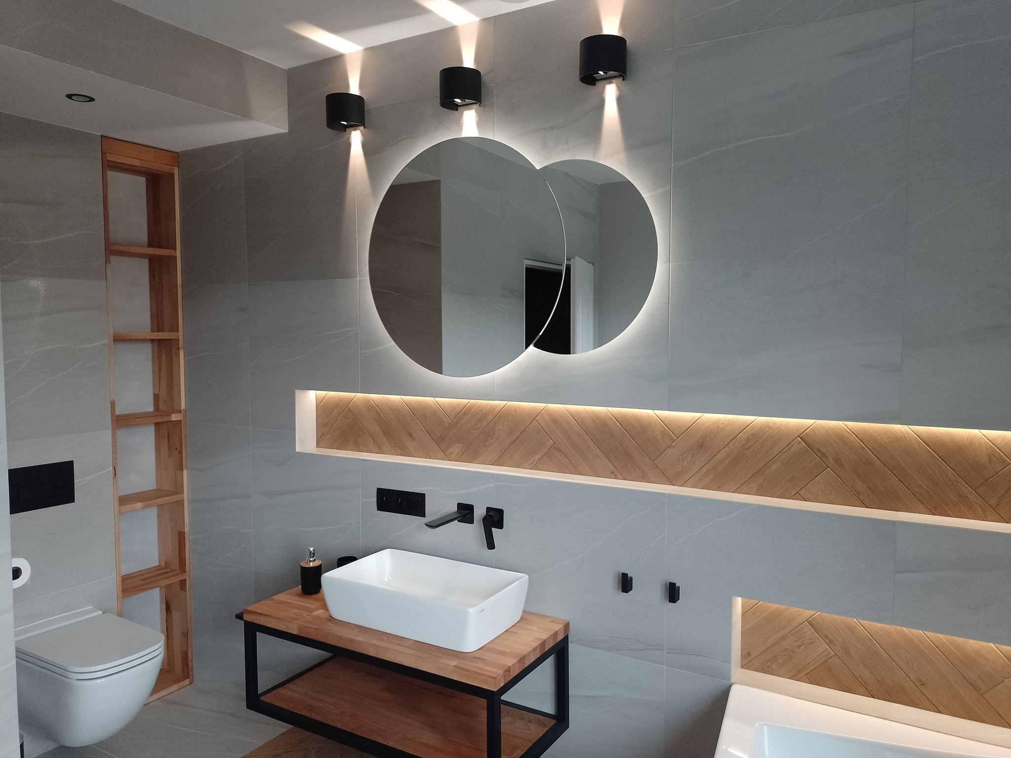 Nowoczesna łazienka z szarymi płytkami, podwójnym okrągłym lustrem z podświetleniem LED, drewnianymi akcentami w układzie jodełki, białą umywalką nablatową na drewnianym stelażu i czarną armaturą.