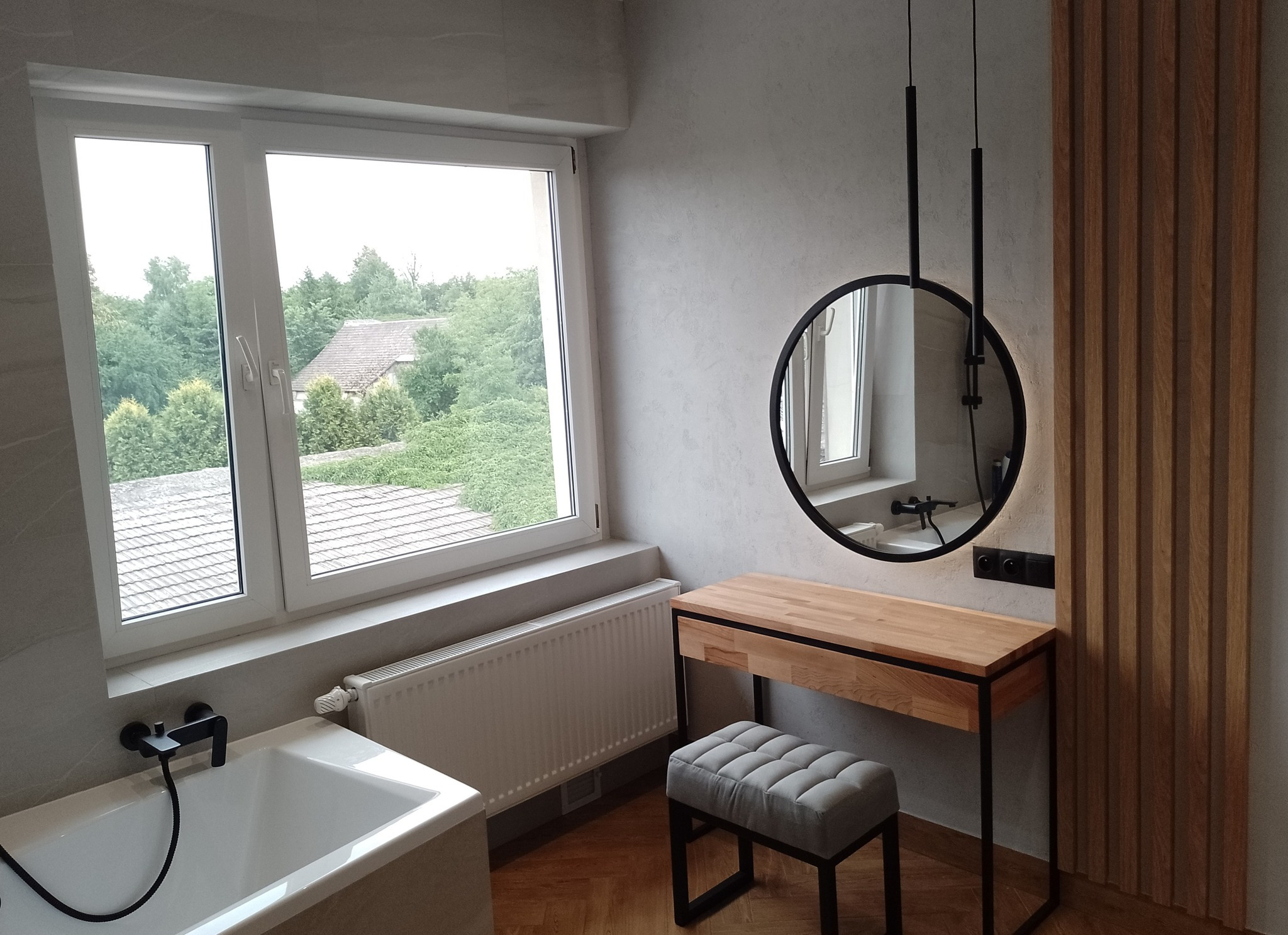 Jasna łazienka z drewnianymi akcentami: okrągłe lustro wiszące nad toaletką z jasnego drewna, szara pufa, biała wanna pod oknem z widokiem na zieleń, dekoracyjny panel ścienny z drewnianych listew.