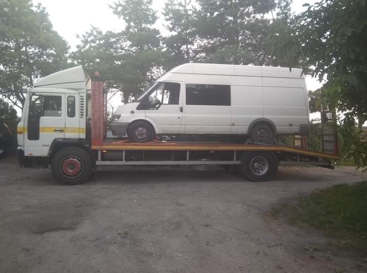 Biały dostawczy samochód na platformie lawety ciężarowej, widok z boku, drzewa w tle.