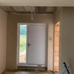 Wnętrze w trakcie prac wykończeniowych: ściany częściowo pokryte jasnym tynkiem, surowy betonowy sufit, białe drzwi wejściowe z widokiem na zewnątrz, fragment muru z cegieł.