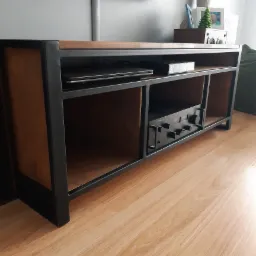 Stolik RTV z drewnianym blatem i czarną metalową ramą, z półkami na sprzęt audio, na podłodze z paneli.