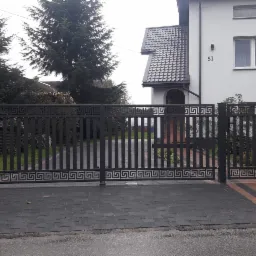 Nowoczesna, metalowa brama wjazdowa w kolorze grafitowym, zdobiona motywem greckim, prowadząca do domu z numerem 51, ułożona na ciemnej kostce brukowej z elementami w kolorze ceglastym.