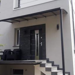 Ranama Inox - Zadaszenie nad wejściem do domu z czarną ramą i przezroczystym wypełnieniem, nowoczesne drzwi wejściowe z elementami stali nierdzewnej, elewacja z szarego kamienia i jasnego tynku, schody wejściowe...