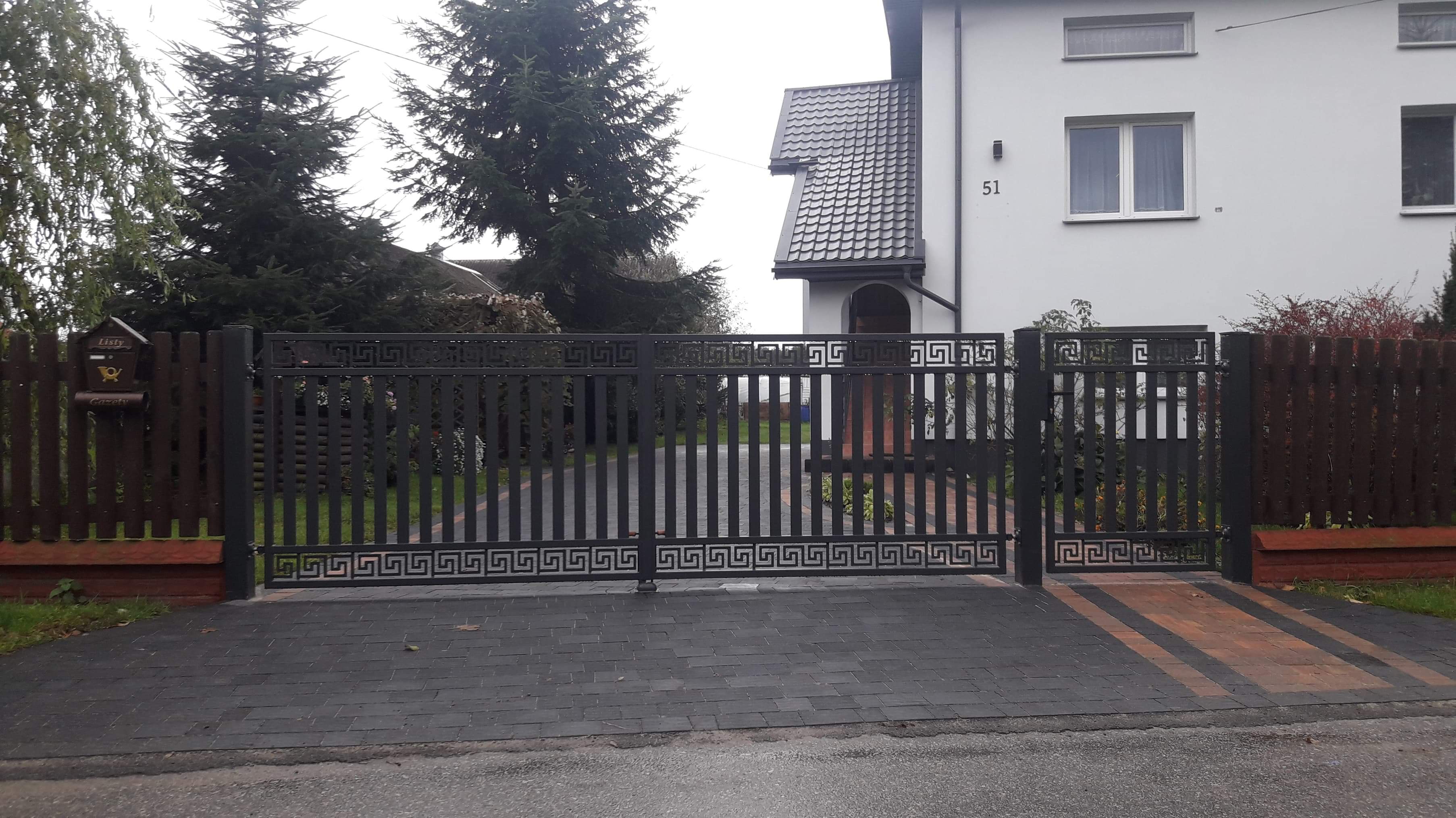 Nowoczesna, metalowa brama wjazdowa w kolorze grafitowym, zdobiona motywem greckim, prowadząca do domu z numerem 51, ułożona na ciemnej kostce brukowej z elementami w kolorze ceglastym.