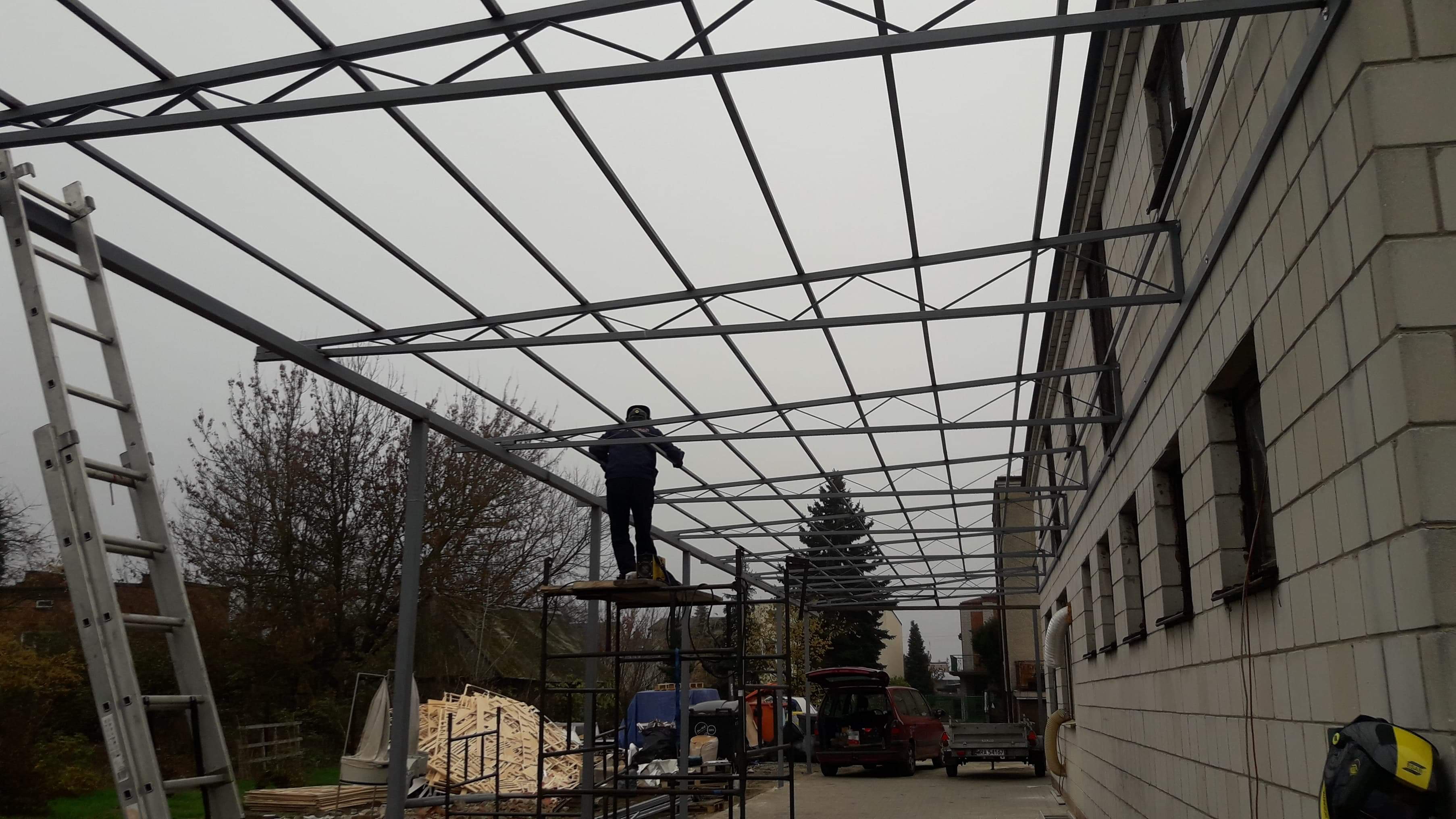 Montaż stalowej konstrukcji zadaszenia przy budynku z pustaków, pracownik na rusztowaniu, drabina oparta o konstrukcję, pochmurne niebo w tle.