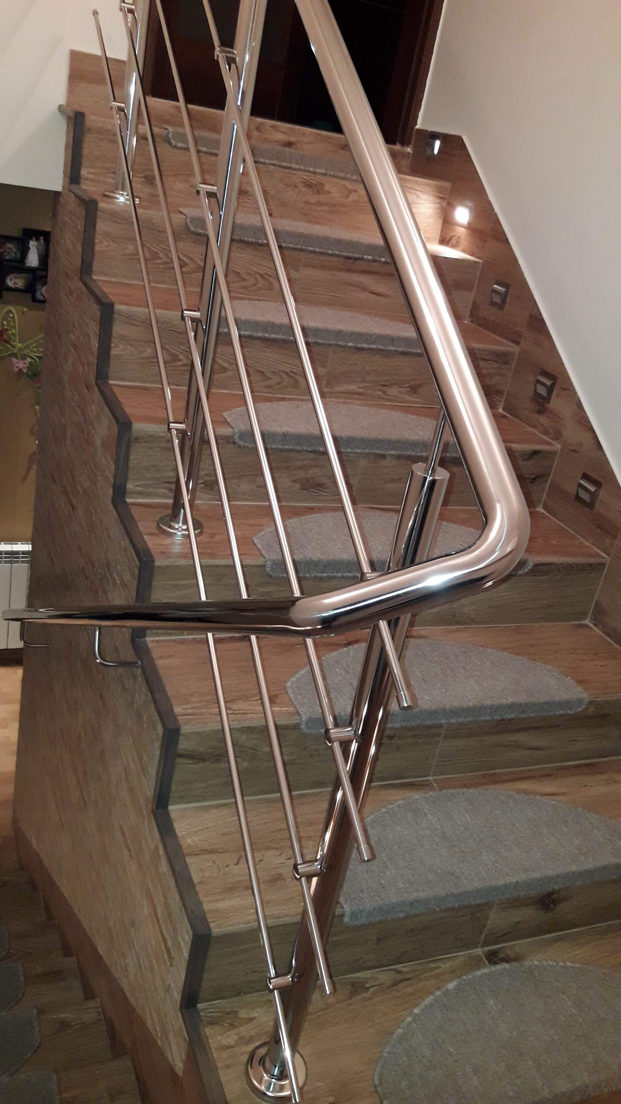 Nowoczesna balustrada ze stali nierdzewnej, z rur o okrągłym przekroju, zamontowana na schodach wyłożonych płytkami imitującymi drewno z naklejonymi półokrągłymi dywanikami.