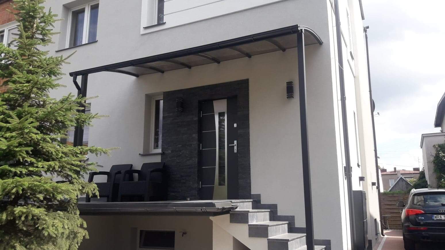 Zadaszenie nad wejściem do domu z czarną ramą i przezroczystym wypełnieniem, nowoczesne drzwi wejściowe z elementami stali nierdzewnej, elewacja z szarego kamienia i jasnego tynku, schody wejściowe...
