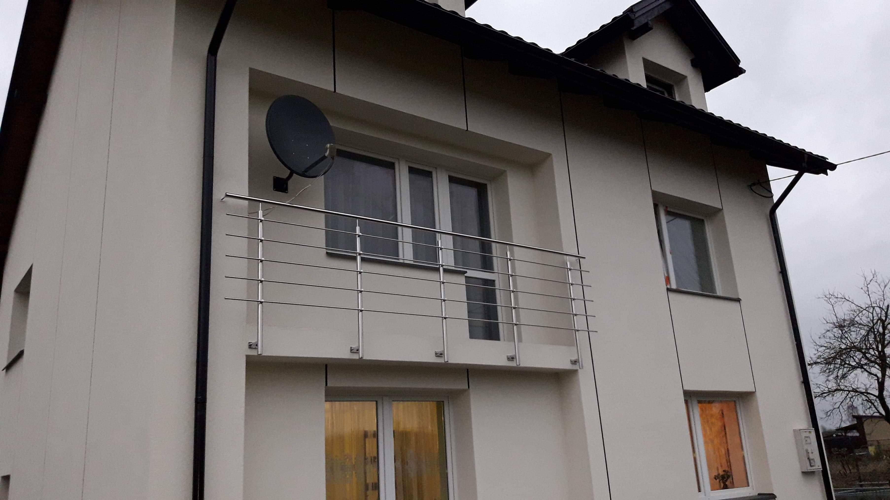 Nowoczesny dom z białymi ścianami i balkonem z nierdzewną balustradą, na balkonie antena satelitarna, widok z dołu w pochmurny dzień.