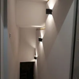 Klatka schodowa z trzema nowoczesnymi czarnymi lampami ściennymi, każda emitująca dwa strumienie światła w górę i w dół, oświetlająca białe ściany i schody prowadzące w dół.