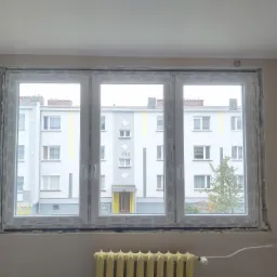 Nowe, trzyskrzydłowe okno PCV zamontowane w otworze okiennym, widoczne szpary wypełnione pianką montażową, na zewnątrz widok na blok mieszkalny.