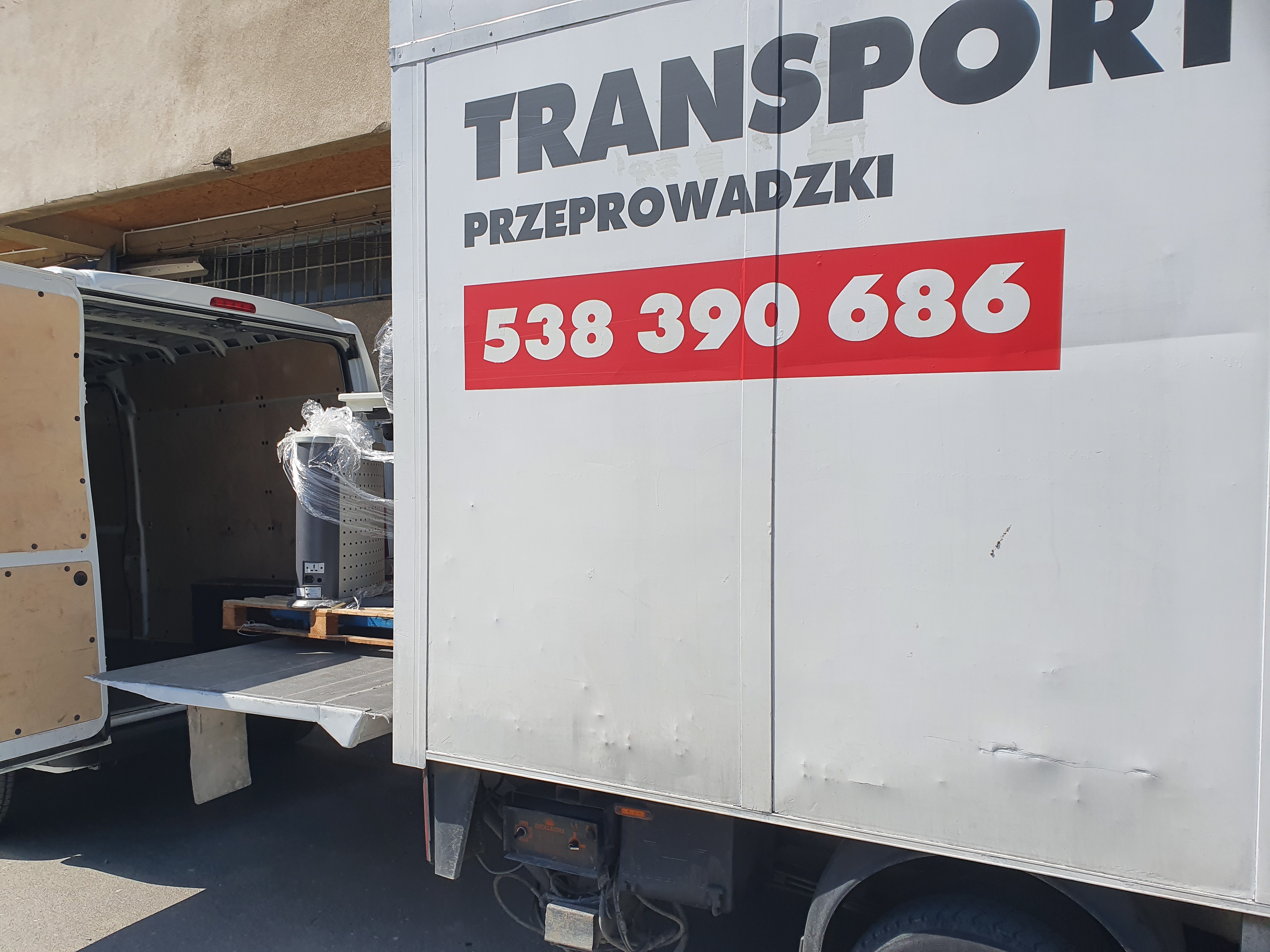Biała furgonetka z otwartymi drzwiami załadowana sprzętem owiniętym folią stretch na palecie; na boku furgonetki widoczne napisy TRANSPORT i numer telefonu.