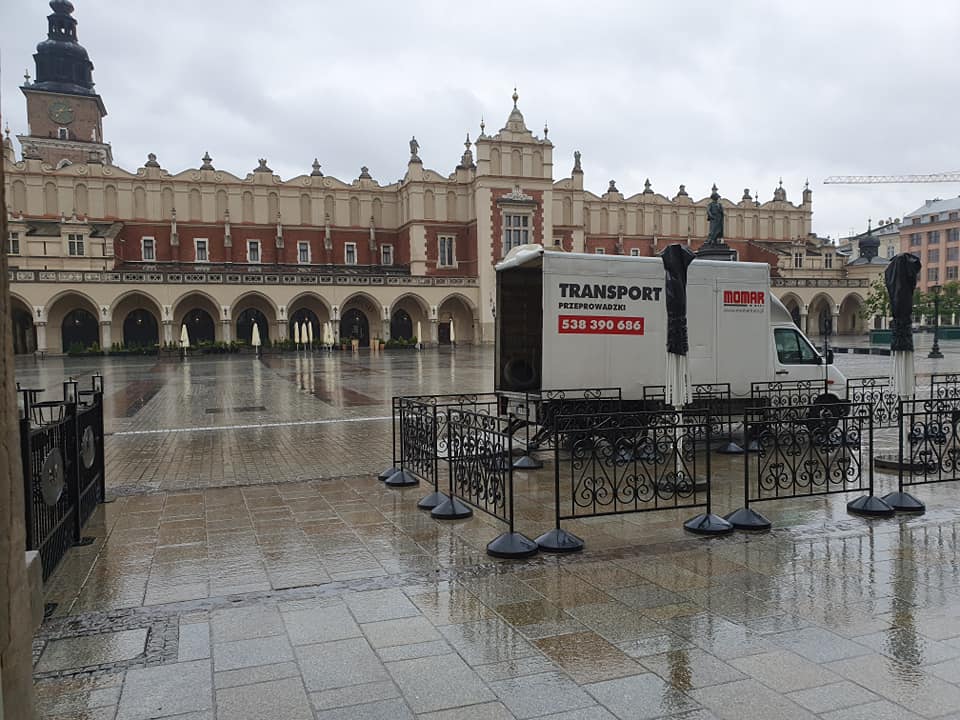 Biała furgonetka z napisem 'Transport Przeprowadzki' i numerem telefonu zaparkowana na mokrym Rynku Głównym w Krakowie, widoczny Sukiennice w tle.