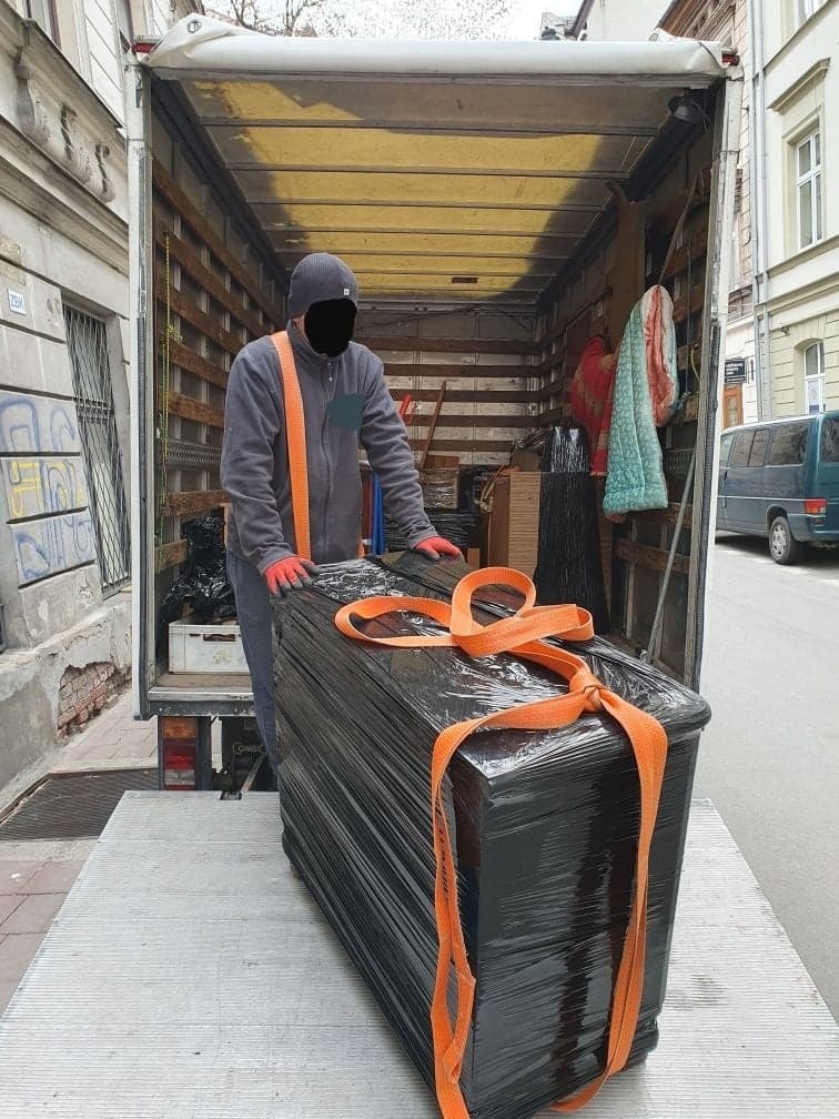 Pracownik w szarym ubraniu i czerwonych rękawicach zabezpiecza czarny mebel owinięty folią stretch pomarańczowymi pasami transportowymi na platformie załadunkowej busa na tle miejskiej ulicy.