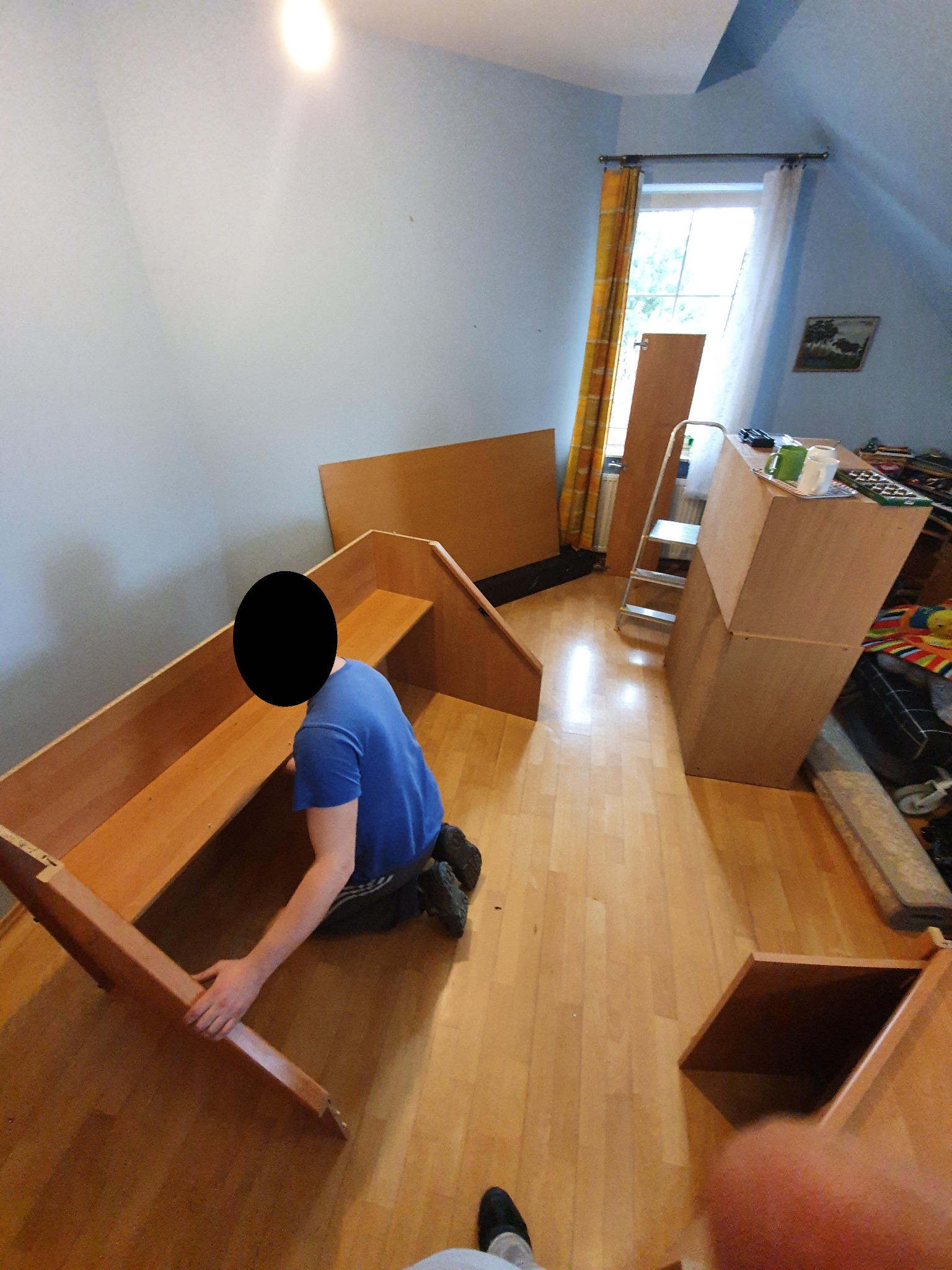 Mężczyzna w niebieskiej koszulce montuje drewniane łóżko piętrowe w pokoju na poddaszu z oknem i żółtą zasłoną, widoczne elementy mebli i drabina.