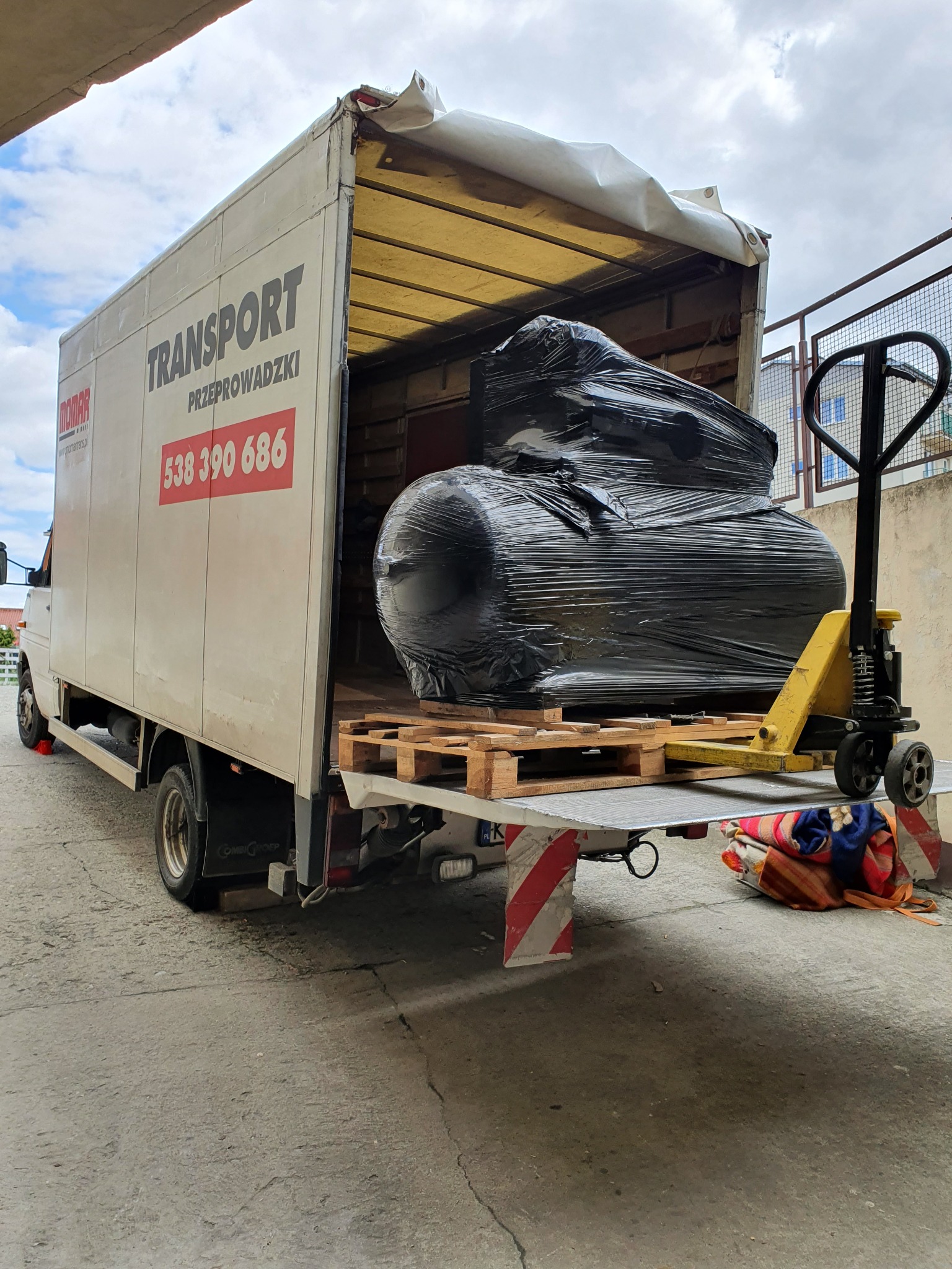 Czarny, owinięty folią ładunek na paletach, transportowany w otwartej przestrzeni ładunkowej białego busa z rampą załadowczą, obok wózek paletowy.