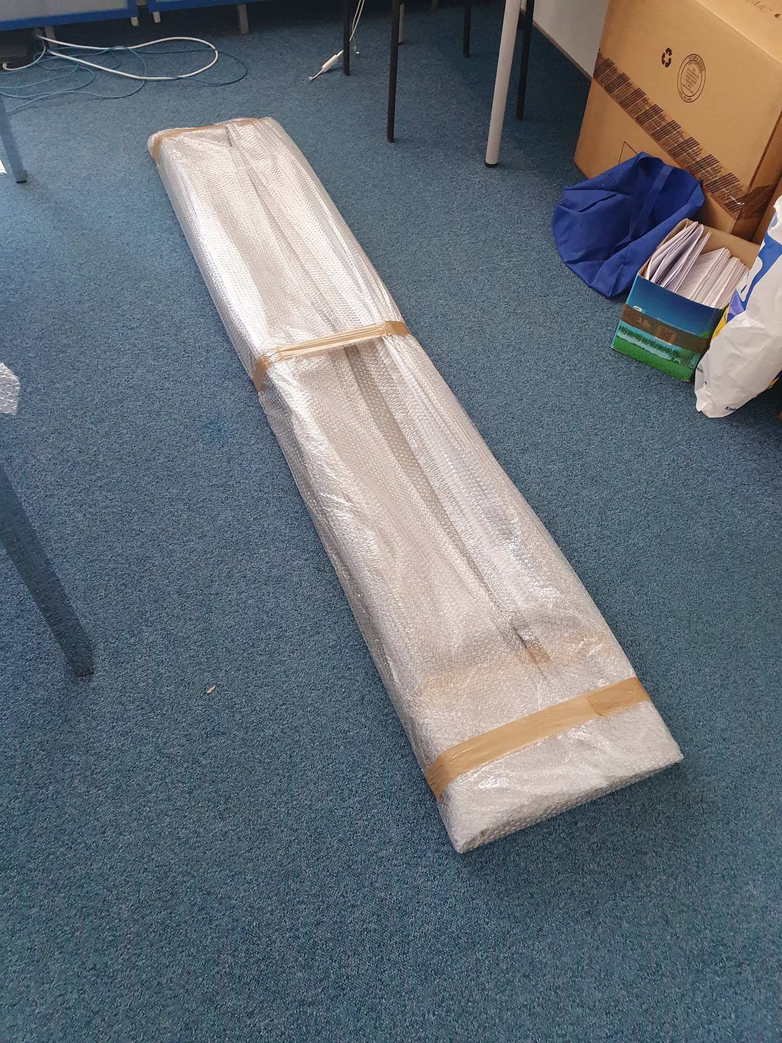 Długa, wąska paczka owinięta folią bąbelkową i zabezpieczona taśmą leży na niebieskiej wykładzinie, gotowa do transportu.