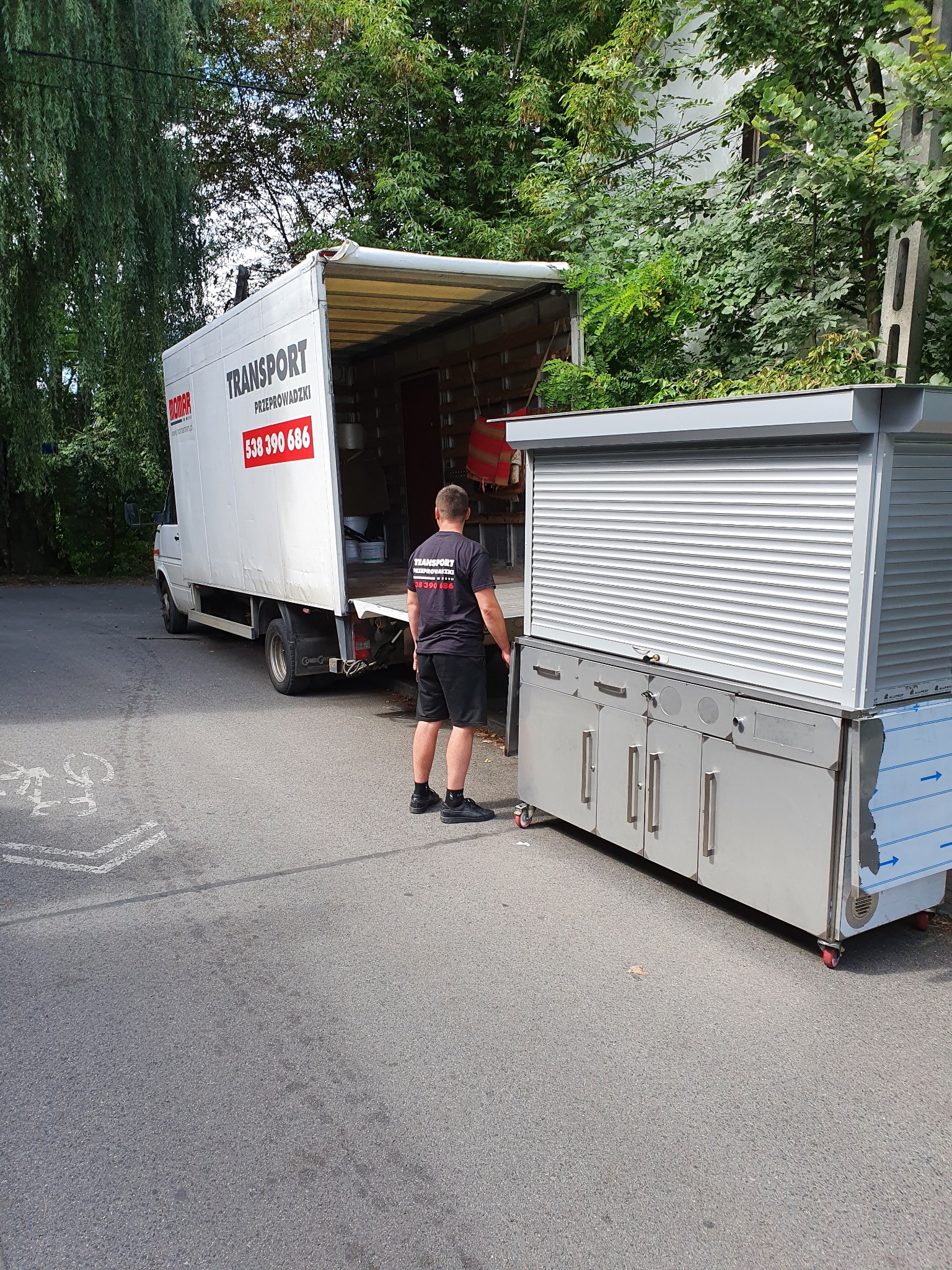 Mężczyzna w czarnej koszulce z logo firmy transportowej stoi obok srebrnej szafki z roletą na kółkach, którą wyładowuje z białego samochodu dostawczego z otwartym tyłem na ulicy.