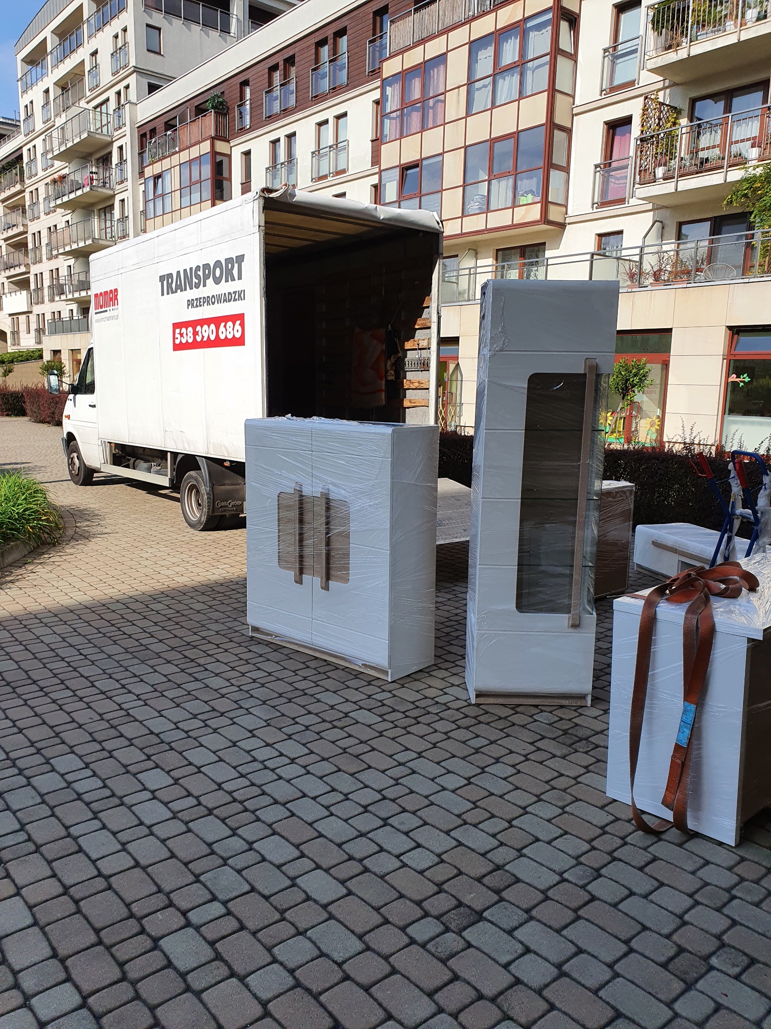 Biała ciężarówka z napisem 'TRANSPORT Przeprowadzki' i numerem telefonu, zaparkowana na brukowanym podjeździe przed budynkiem mieszkalnym, z zapakowanymi w folię meblami gotowymi do załadunku.