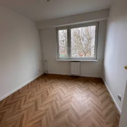 Pusty pokój po remoncie z parkietem ułożonym w jodełkę, białe ściany, okno z widokiem na drzewa bez liści, grzejnik pod oknem, złota klamka przy białych drzwiach.