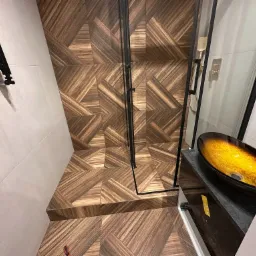 Nowoczesna łazienka z kabiną prysznicową typu walk-in, wykończona płytkami imitującymi drewno ułożonymi w geometryczny wzór, oraz umywalką nablatową w odcieniach bursztynu.