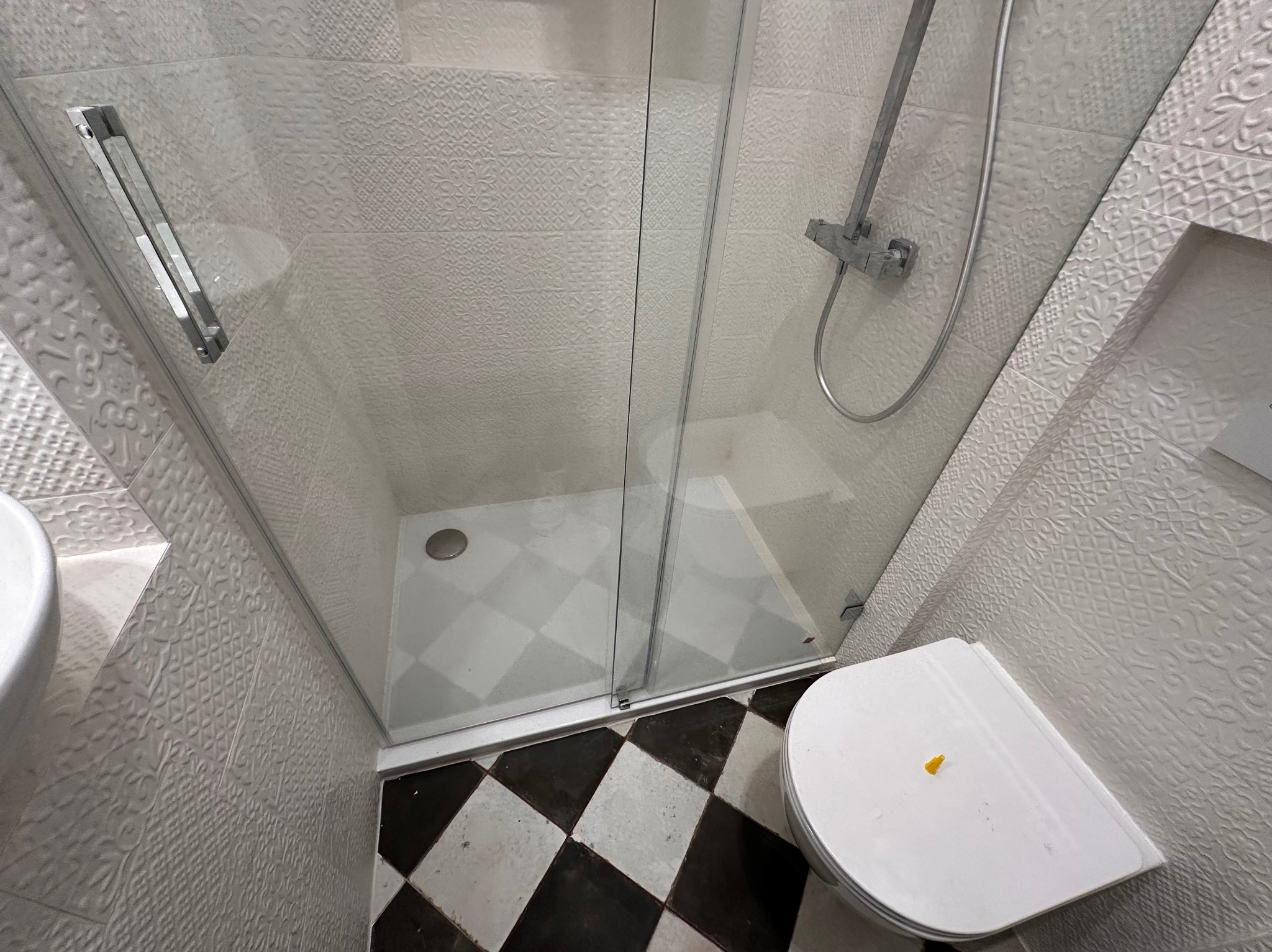 Kabina prysznicowa z deszczownicą i białą, wzorzystą glazurą ścienną, czarno-biała szachownica na podłodze i toaleta z żółtą naklejką.