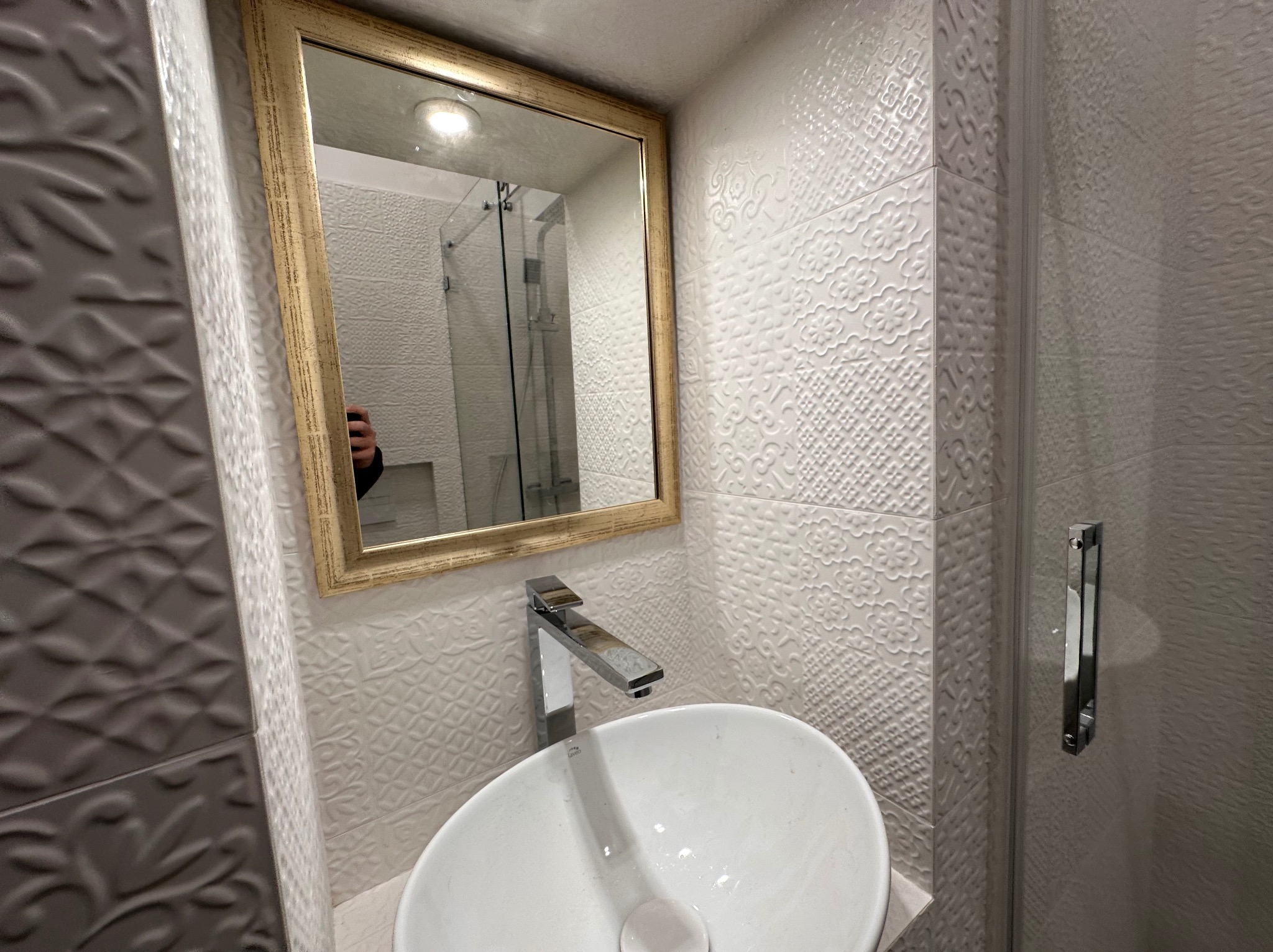 Biała umywalka nablatowa z chromowaną baterią na tle dekoracyjnych, białych płytek ściennych i lustra w złotej ramie, odbijającego kabinę prysznicową.